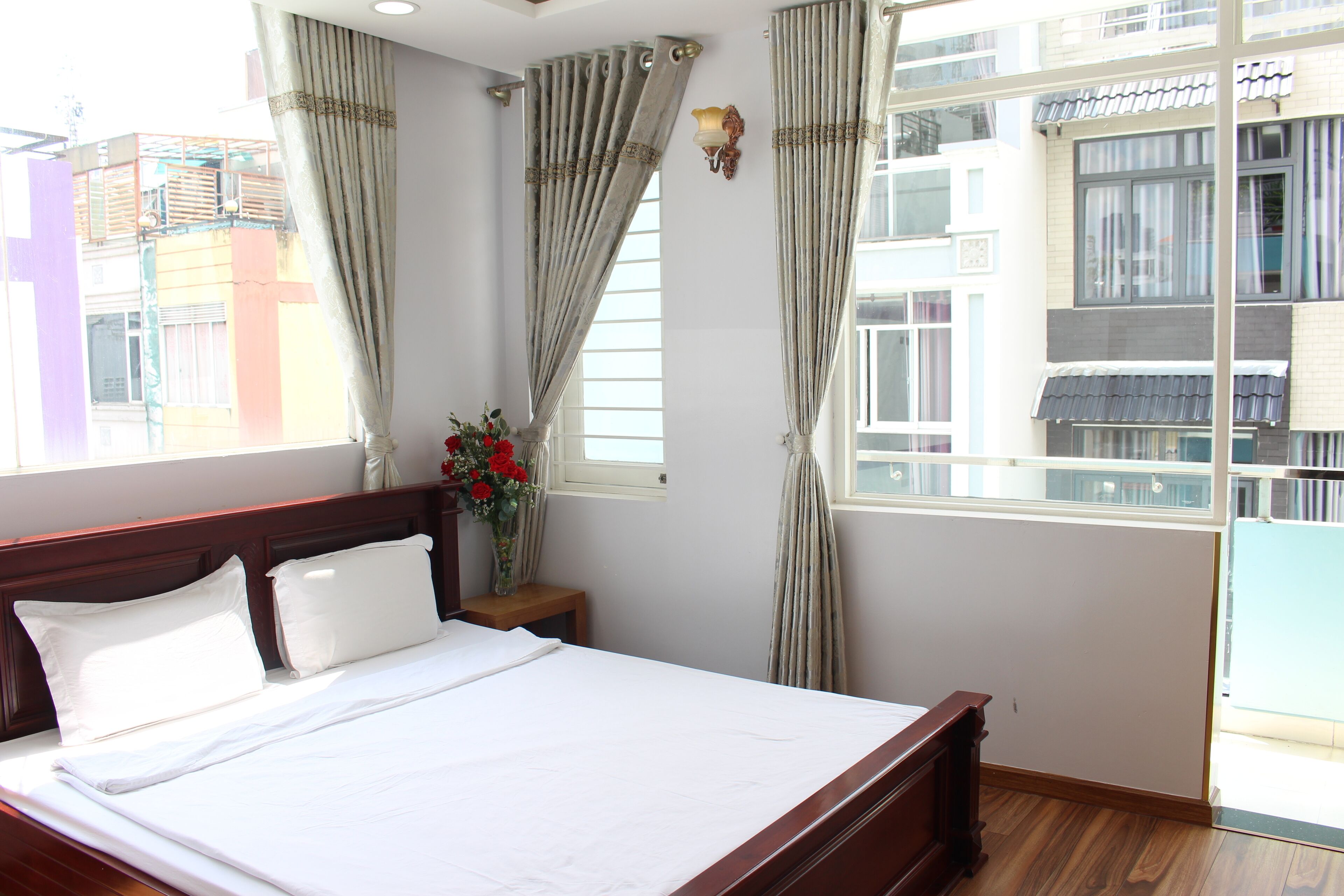 A hotel room with a large bed, curtains, a nightstand with a vase of flowers, and a view of buildings through the window.