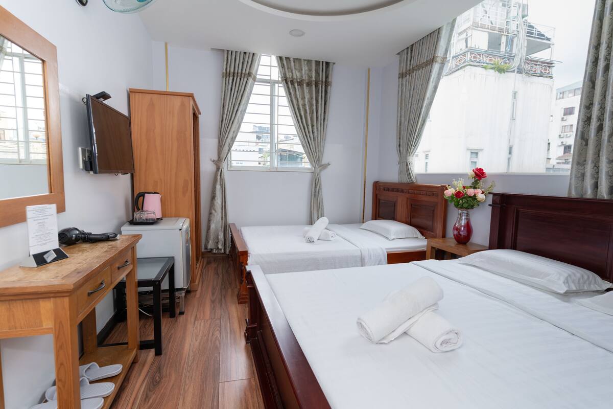 Habitación de hotel con dos camas, un escritorio de madera, una silla, una mesita con un florero y una ventana con cortinas.