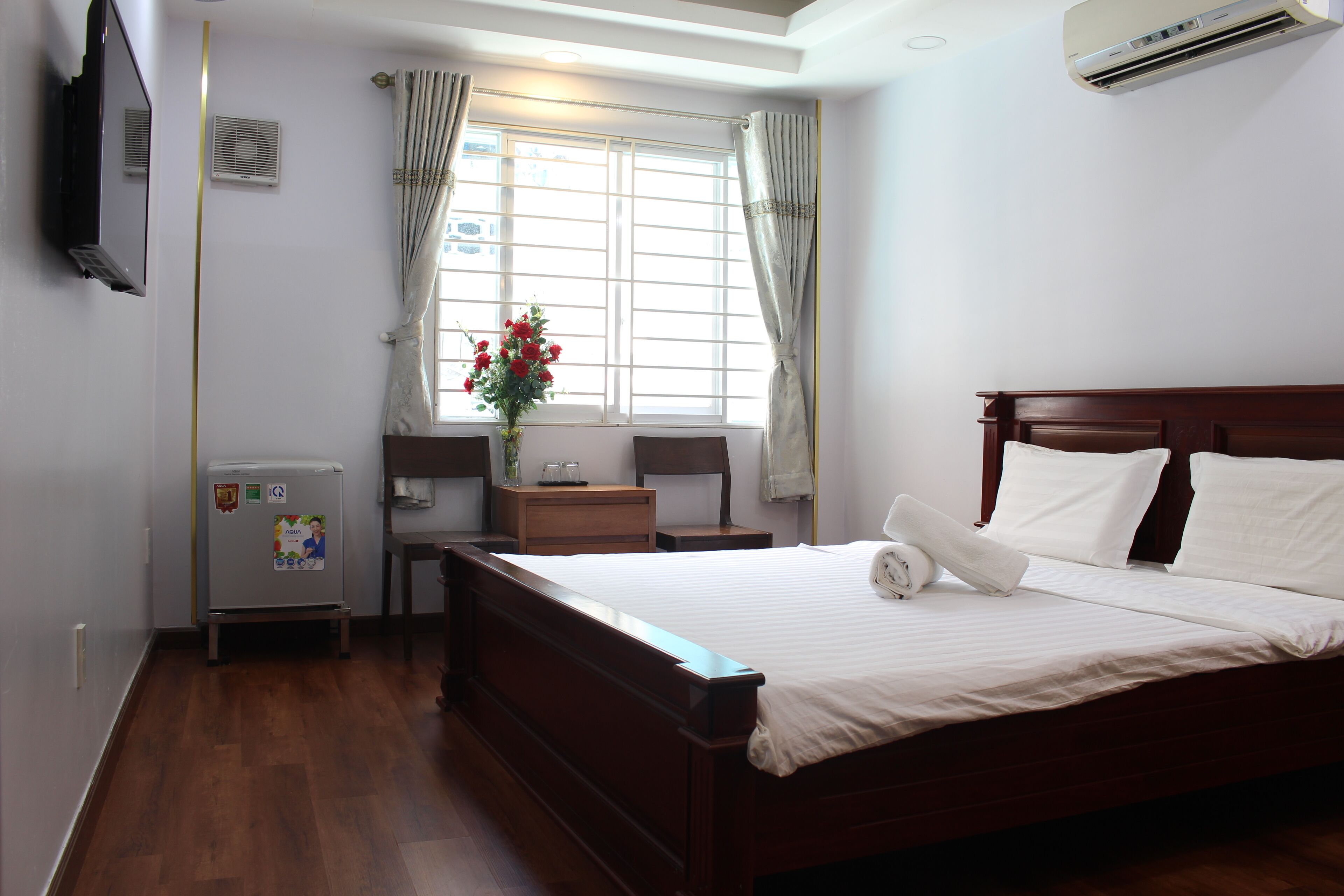 A hotel room with a wooden bed, a desk with a chair, a small refrigerator, a window with curtains, and an air conditioning unit.