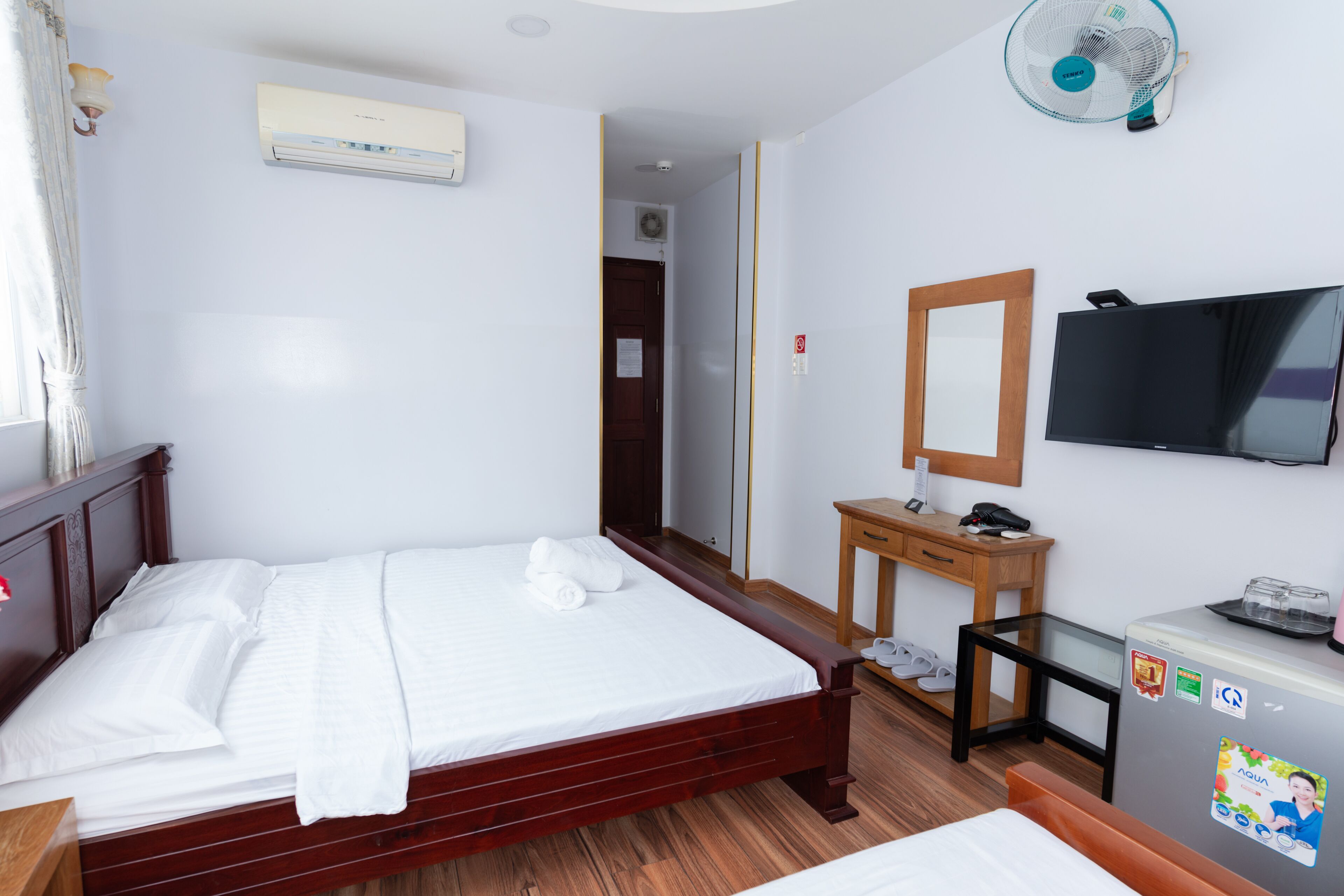 Quarto de hotel com cama de madeira, uma secretária pequena com espelho, televisão e frigorífico.