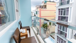 Un balcón con una mesa y sillas de madera, barandilla de vidrio y vista a edificios cercanos.