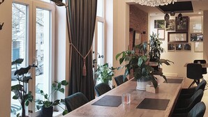 Een ruime eethoek met een grote houten tafel, zwarte stoelen en een kroonluchter. Achtergrond met potplanten en een bakstenen muur.