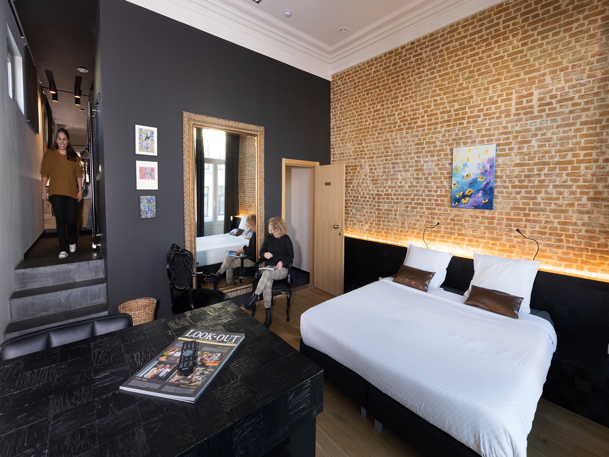 Une chambre d’hôtel moderne avec un grand lit, un coin salon avec un magazine, un miroir et un mur en briques.