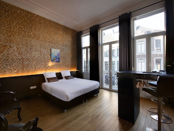A hotel room with a brick wall, a bed, a desk with a chair, and a view of the outside. - Goodnight Antwerp (Antwerp)