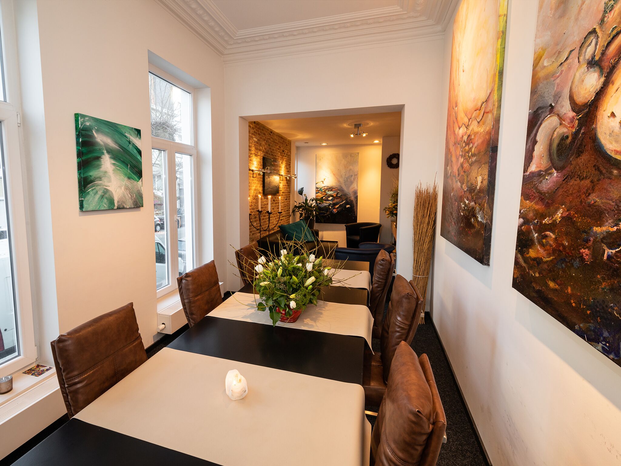 Un coin repas avec une table dressée pour quatre personnes, entourée de fauteuils en cuir marron, un vase rempli de fleurs et des œuvres d’art accrochées aux murs.