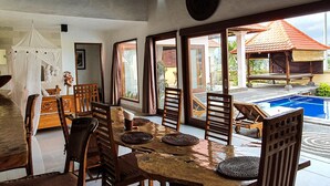 A dining area with a wooden table and chairs, a white canopy, and a view of an outdoor swimming pool. - Villa Tatiapi Ubud (Ubud)