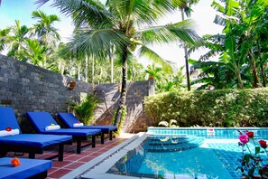Uma área à beira da piscina com espreguiçadeiras, parede de pedra e vegetação tropical.
