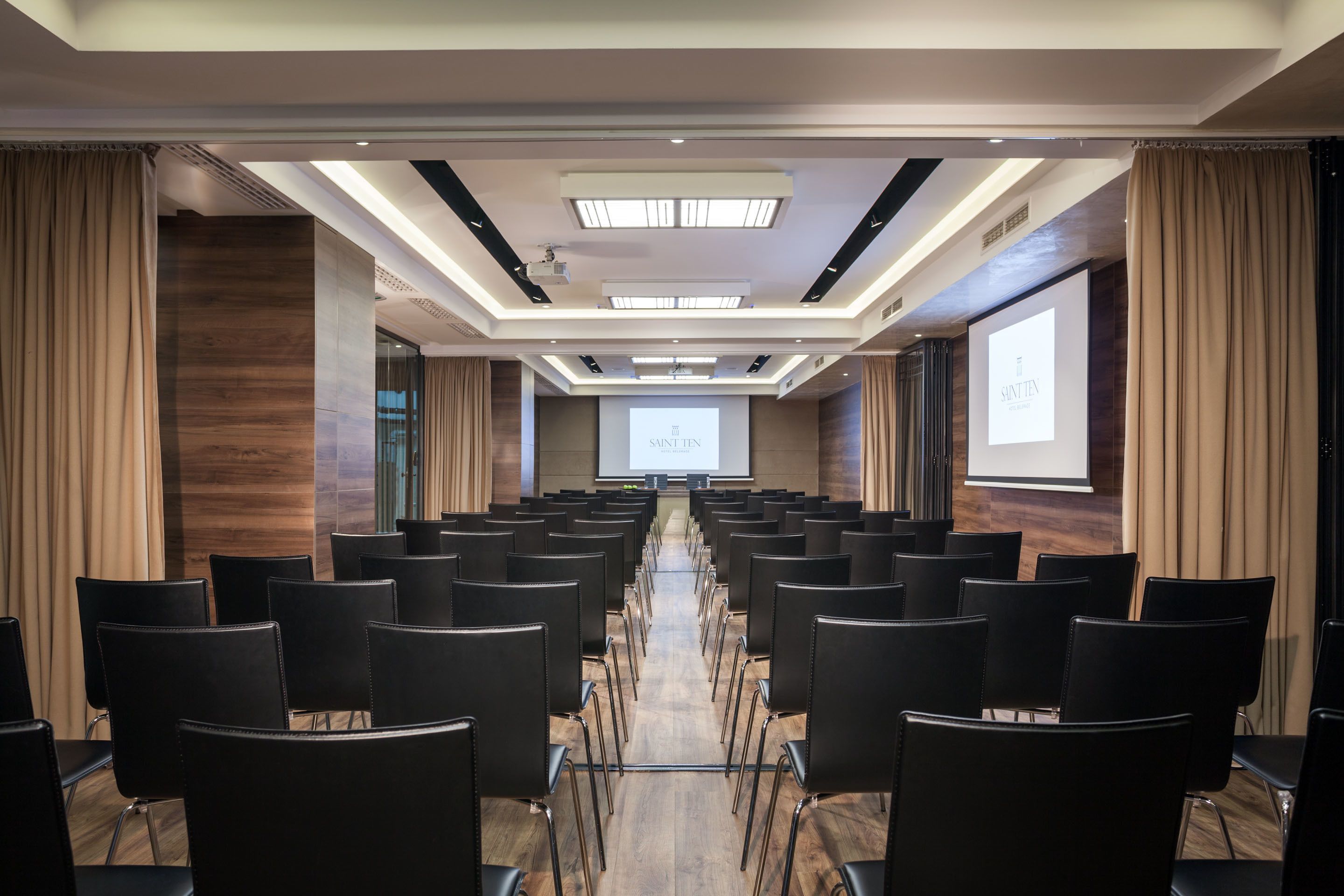 Un salón de conferencias con filas de sillas negras, suelo de madera y una pantalla grande al frente.