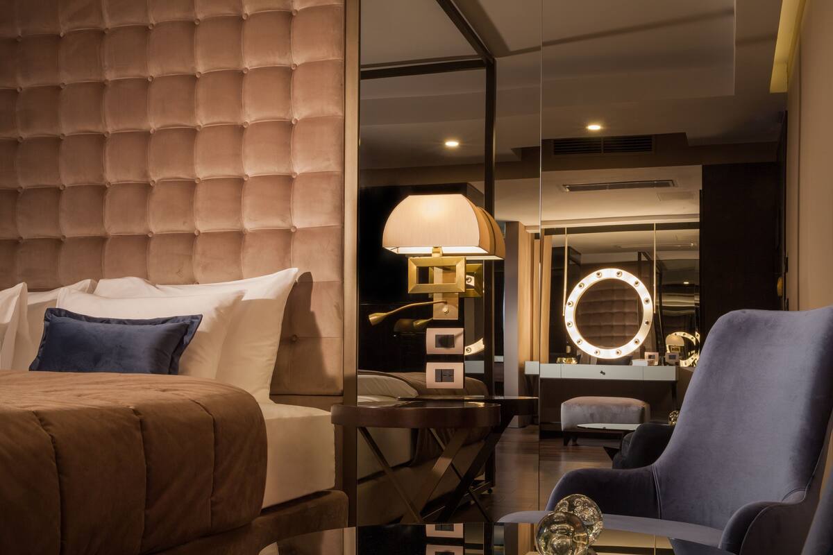 Une chambre d’hôtel moderne avec une tête de lit capitonnée, un grand miroir avec éclairage, une table ronde avec une lampe et un fauteuil bleu.
