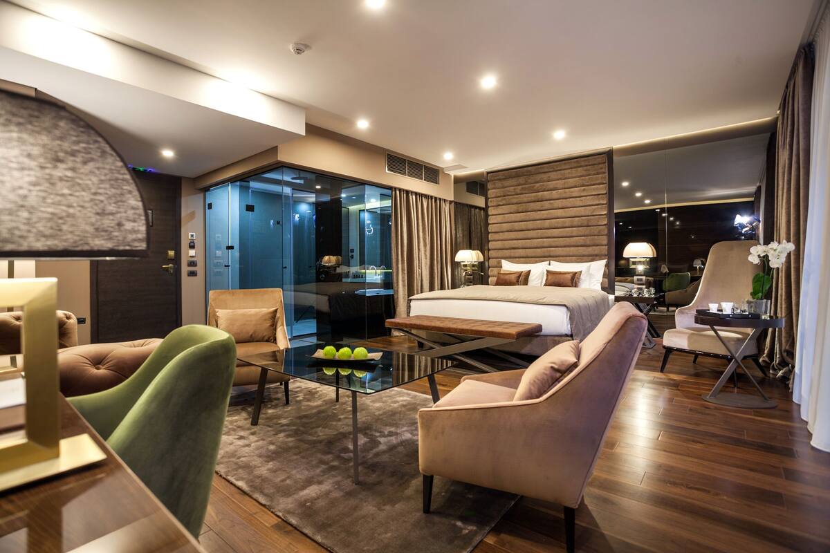 Une chambre d’hôtel moderne dotée d’un grand lit, d’une cloison en verre, d’un coin salon avec des fauteuils et d’une table à manger.