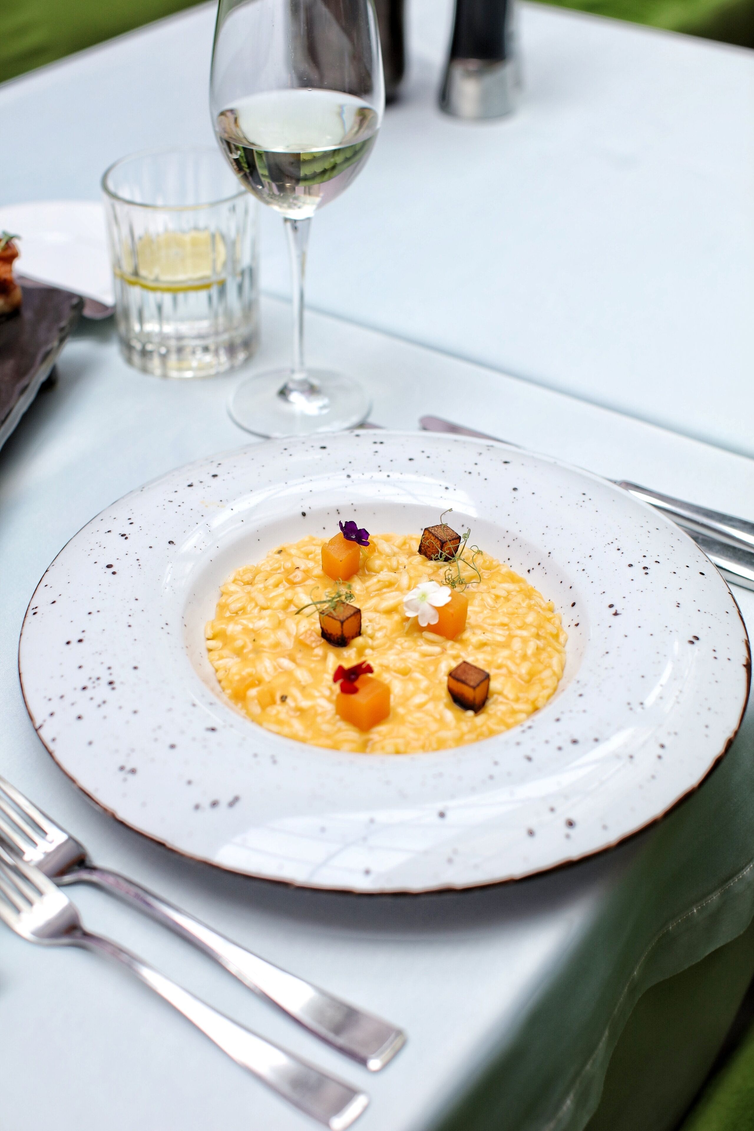 A plate of risotto with garnishes, a glass of white wine, and a small glass of water on a table.