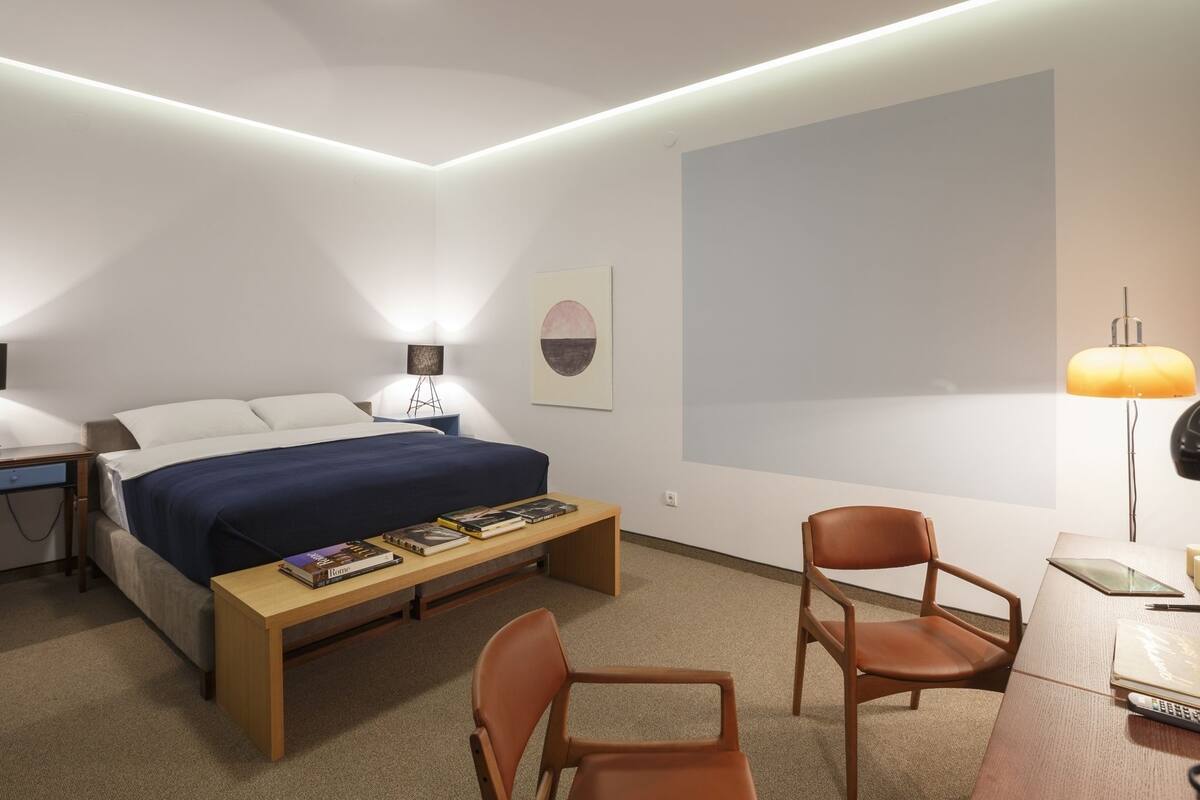 A modern hotel room with a neatly made bed, a desk with a chair, and a bedside table with books.