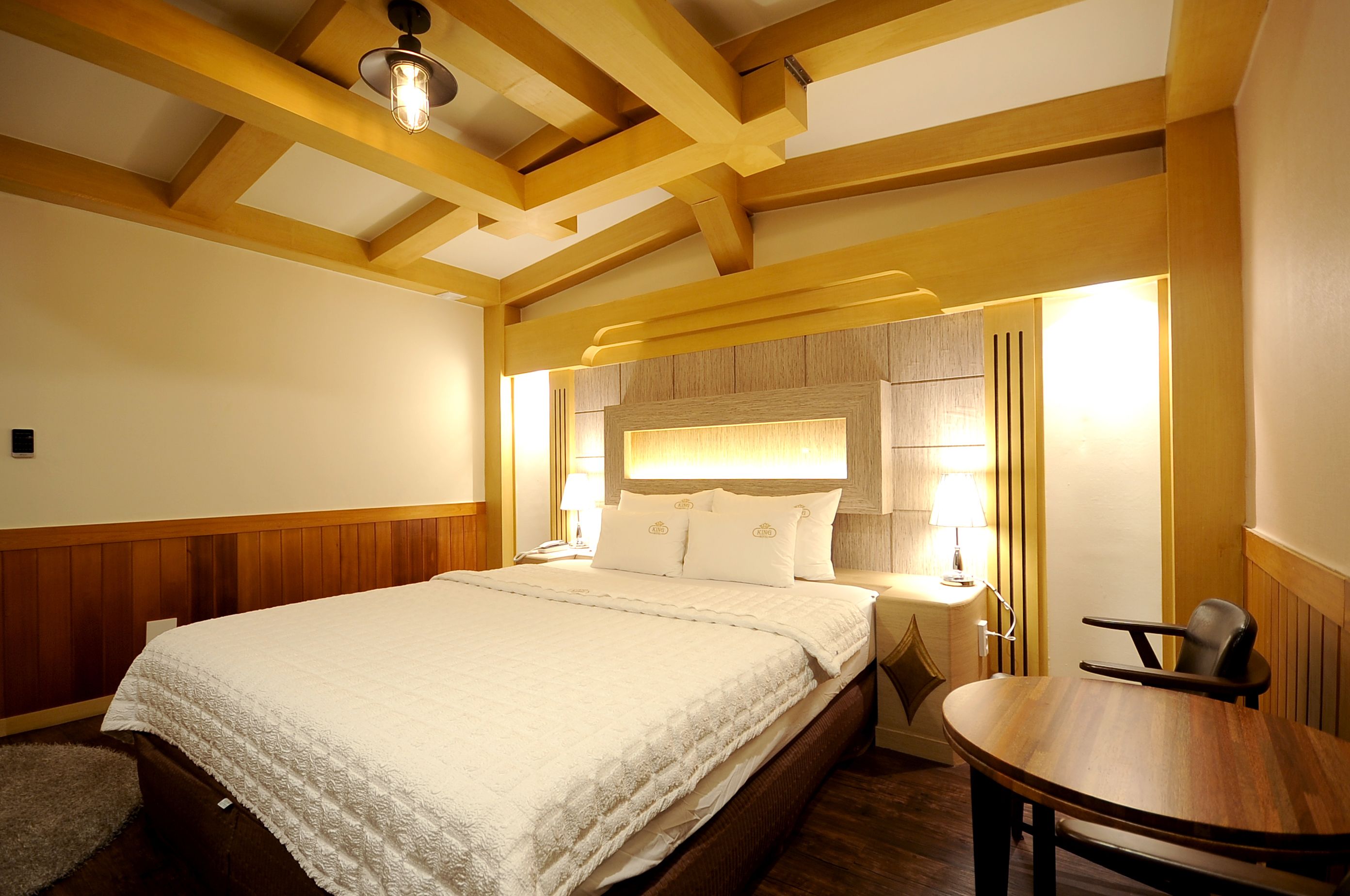 A bedroom with a large bed, wooden ceiling beams, a bedside table with a lamp, and a small round table with a chair.