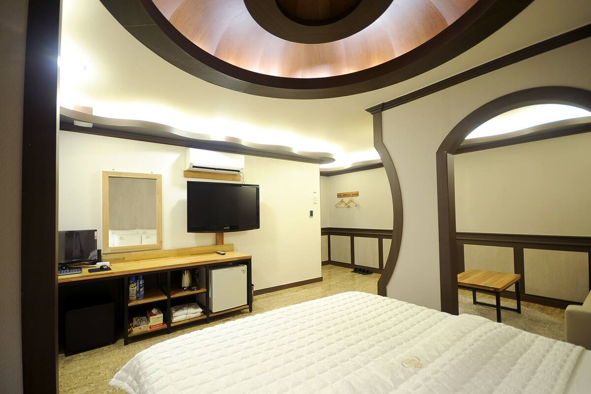 A hotel room with a bed, a TV, a desk with a computer, a small refrigerator, and a shoe rack.