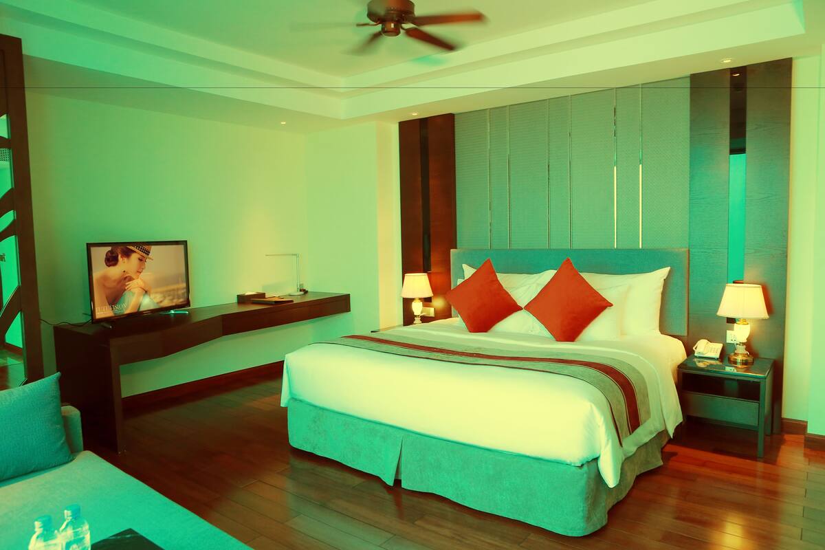 Quarto de hotel moderno com uma cama grande, duas mesas de cabeceira com luminárias, um ventilador de teto e uma TV sobre um suporte.