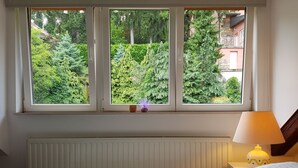 A bedroom with a bed, a nightstand with a lamp, and a window with a view of greenery and buildings. - Taban Hostel Zagreb - hostel (Zagreb)