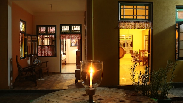 A candle in a glass holder on a wooden table, with a view of a room with a dining table and chairs, and a doorway leading to another room. - Villa Ceylon (Kimbulapitiya- Katunayake)