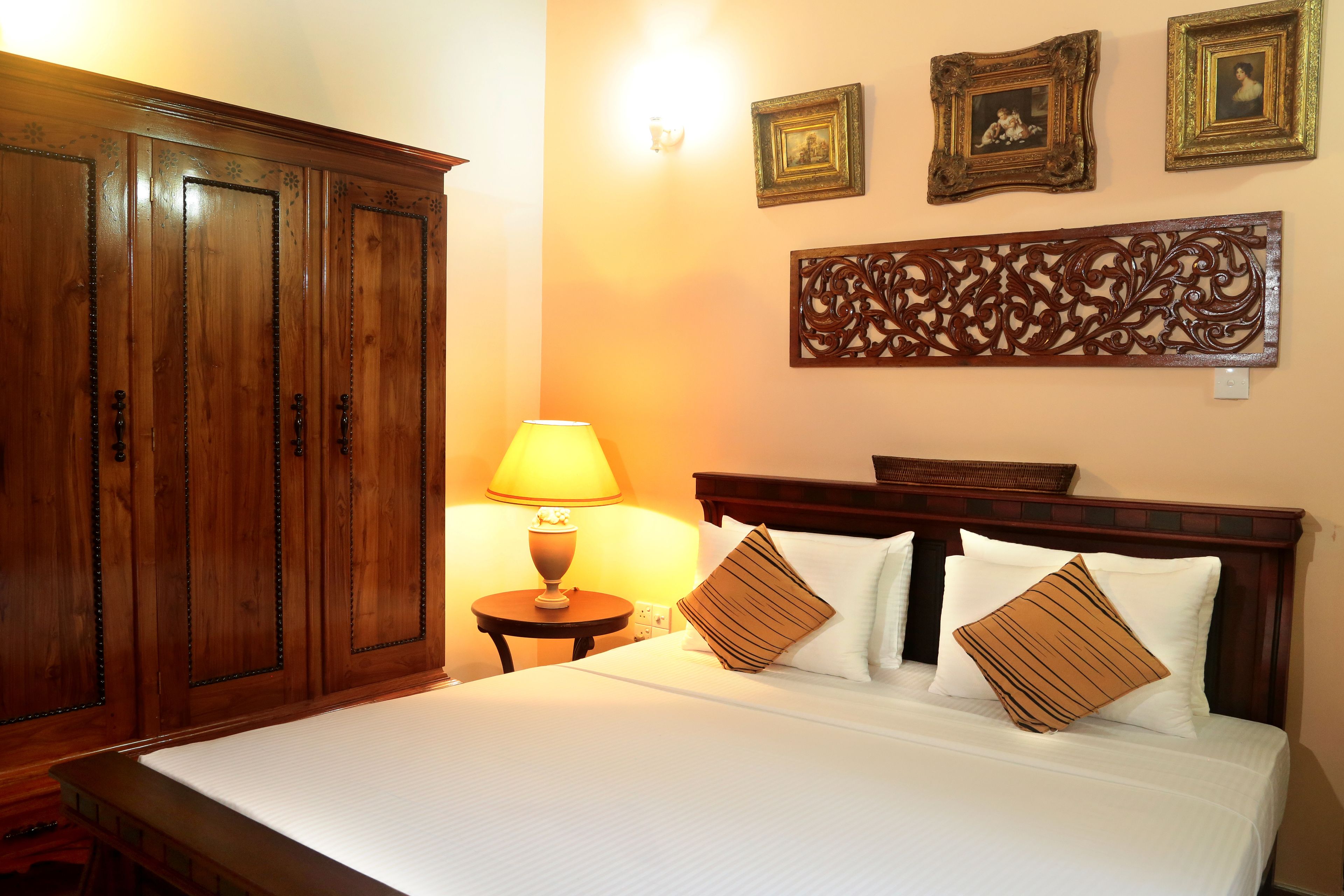 Habitación de hotel con un armario de madera grande, una cama con ropa de cama blanca y almohadas estampadas, una lámpara de noche y decoración en la pared.