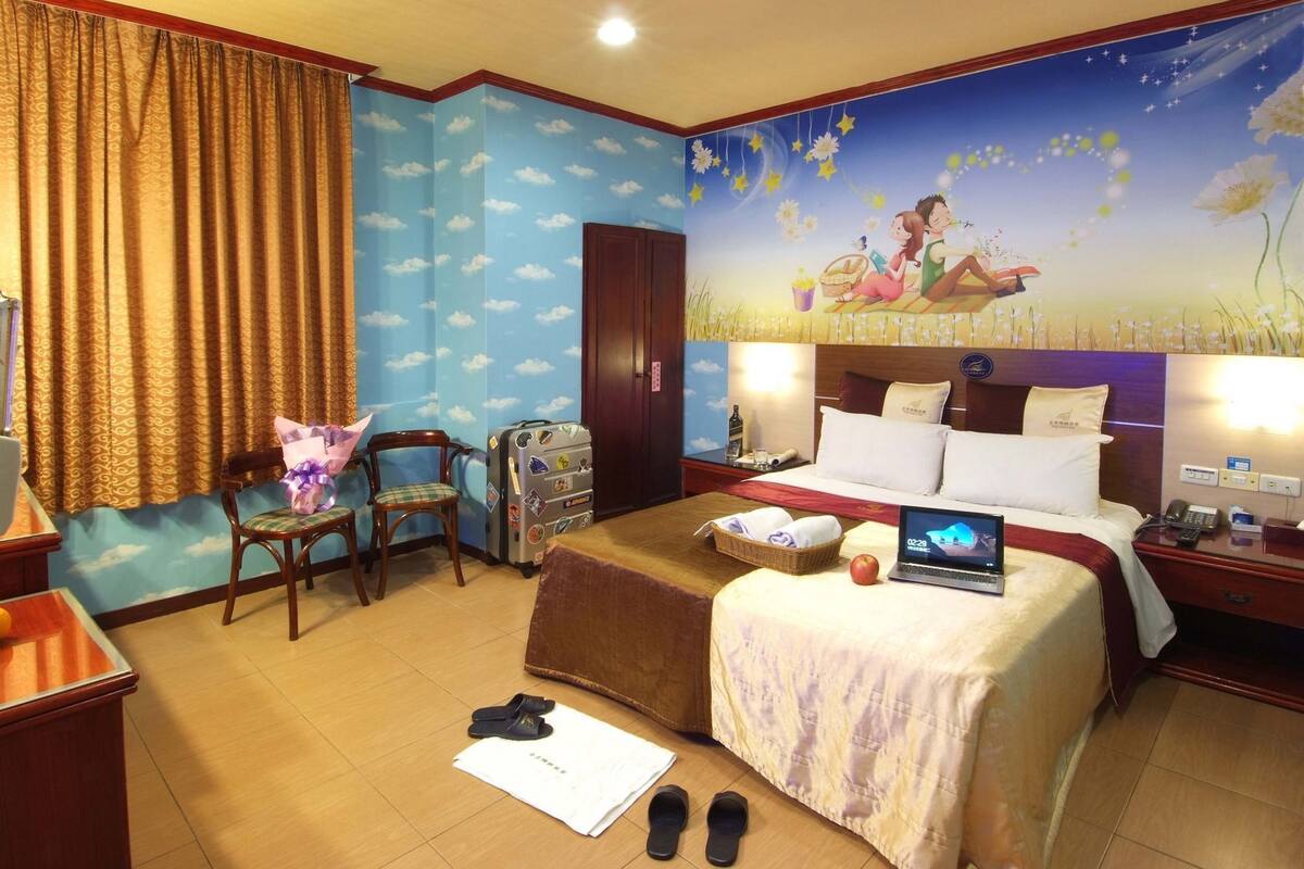 A hotel room with a bed, a desk, a chair, a suitcase, a laptop, slippers, and a painting on the wall.
