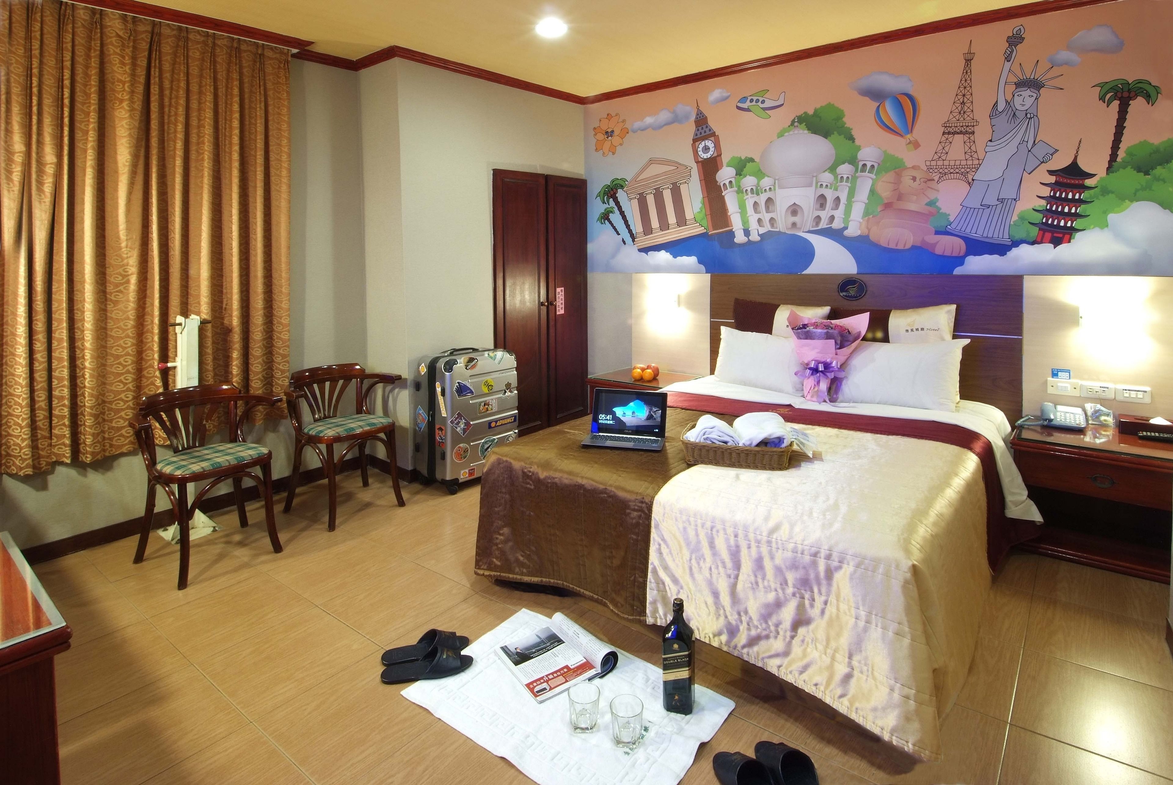 A hotel room with a bed, a desk with a laptop, a chair, a suitcase, and a wall mural.