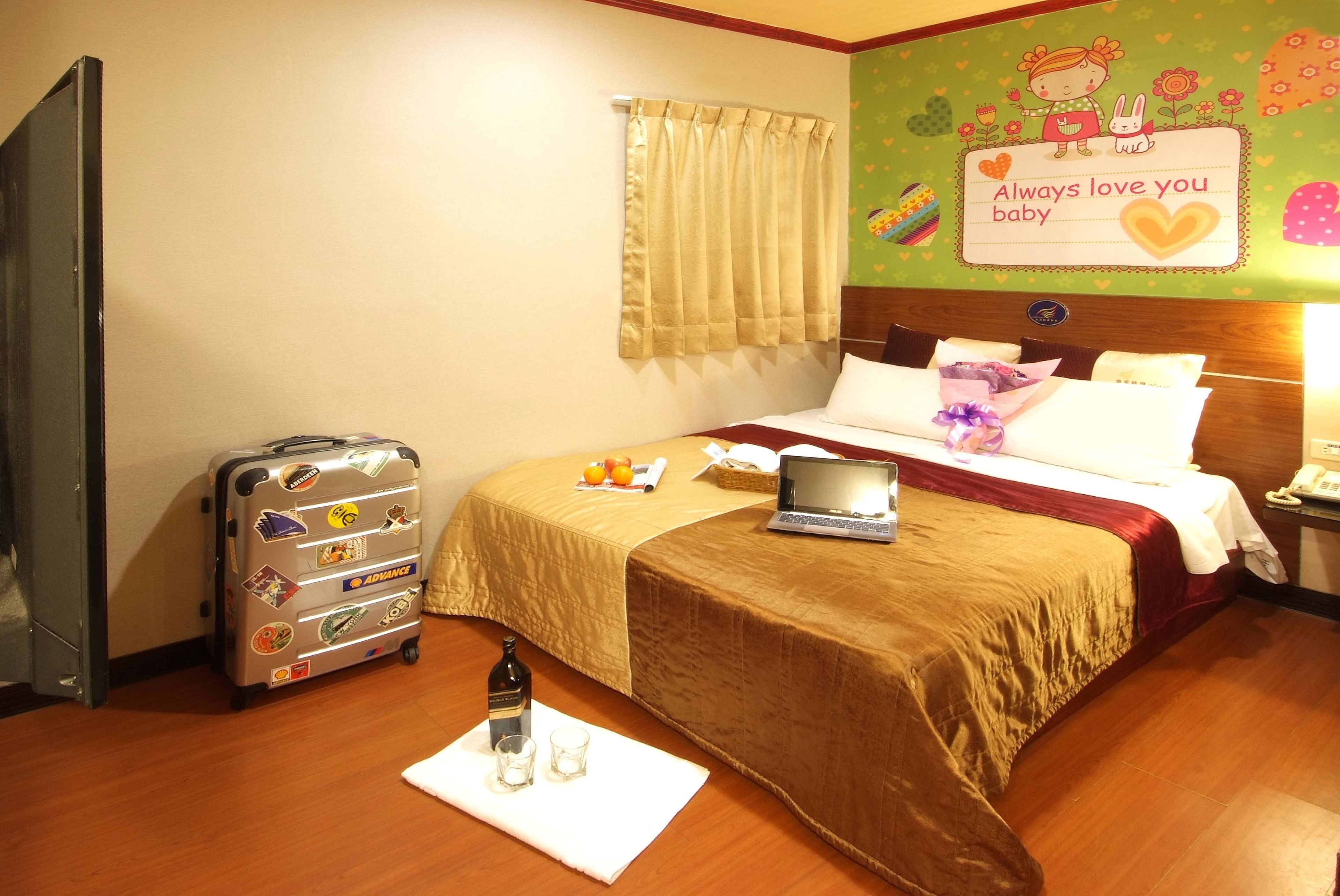 A hotel room with a bed, a laptop, a suitcase, and a bottle of water on a white mat.