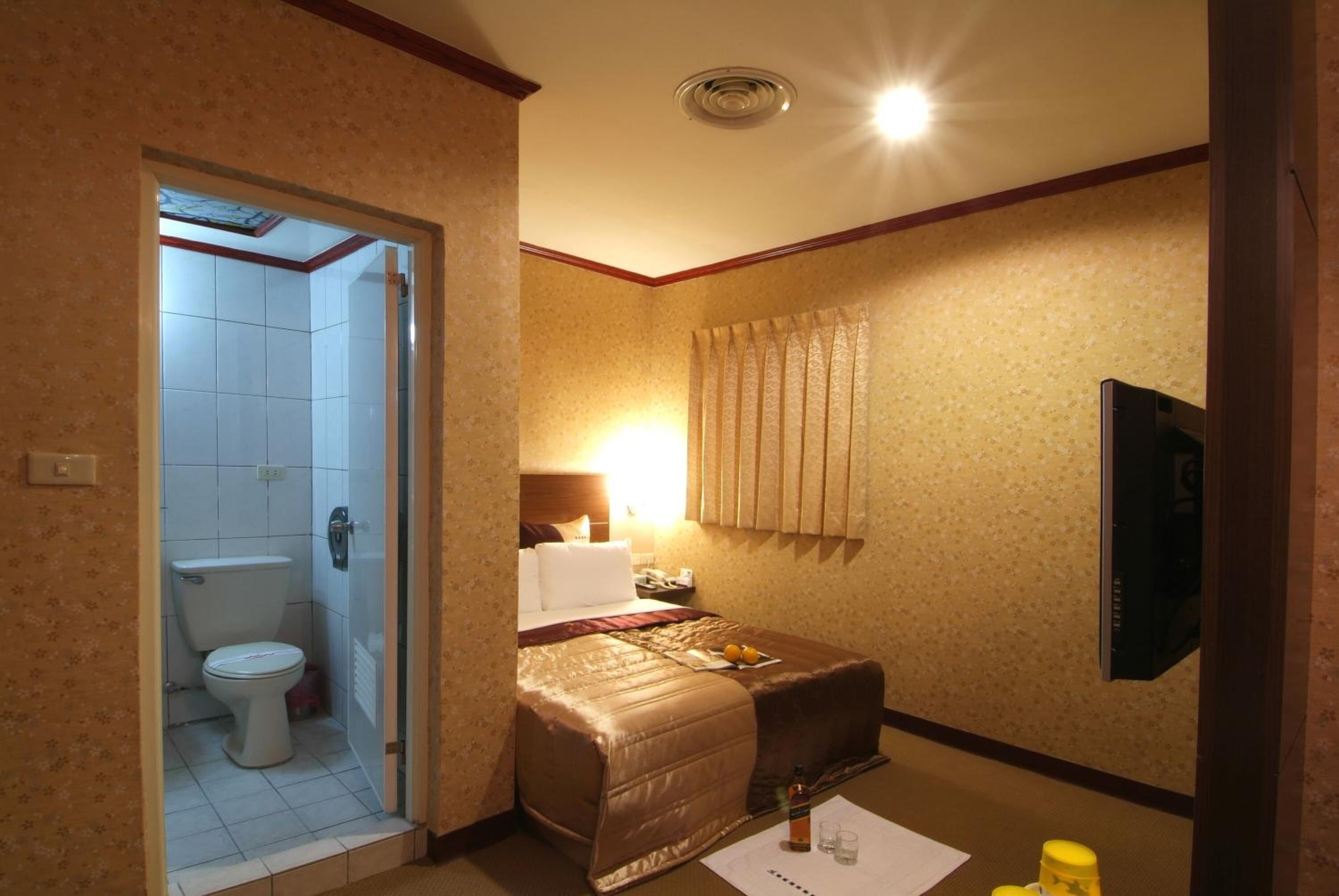 A hotel room with a bathroom, a bed with a brown bedspread, a bedside table with a bottle and glasses, and a TV mounted on the wall.