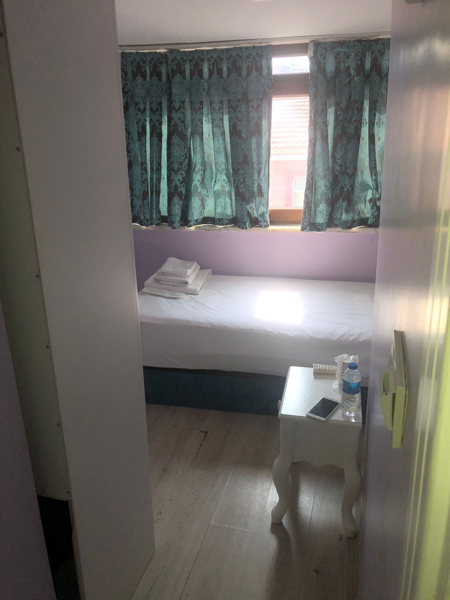 Una habitación pequeña con una cama individual, una mesita con una botella de agua y una ventana con cortinas.