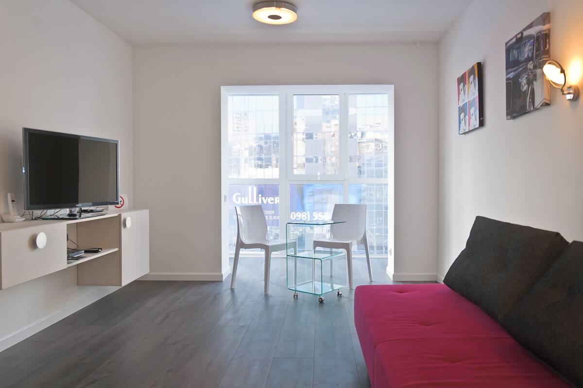 A modern living room with a flat-screen TV, a glass coffee table, two white chairs, a red sofa, and a city view through large windows.