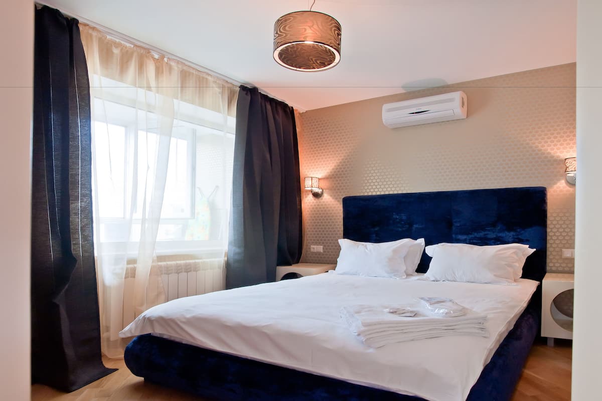 A neatly made bed with white linens, a dark blue headboard, and a wall-mounted air conditioner.