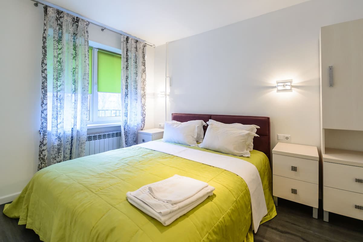 A neatly made bed with a green bedspread and white pillows, a bedside table with a lamp, and a window with floral curtains.