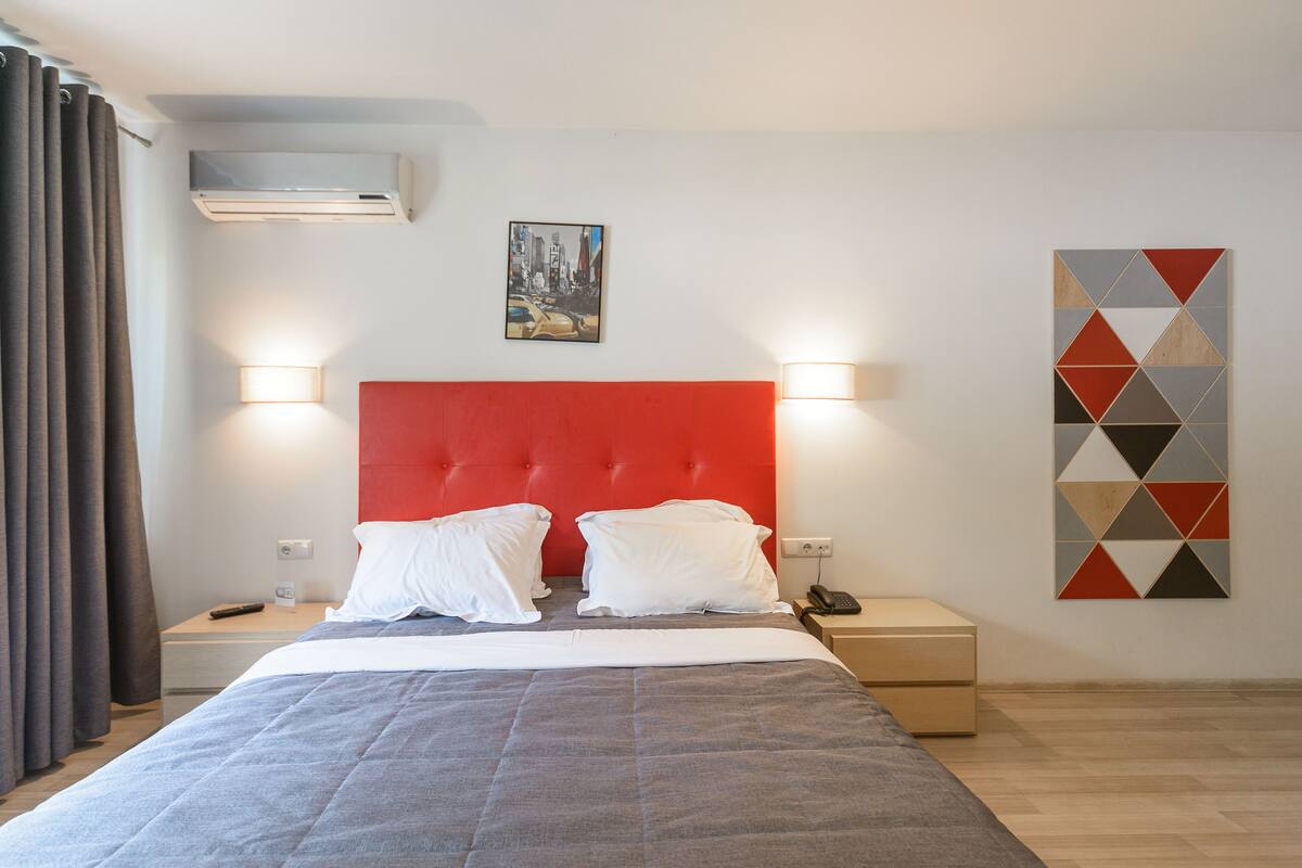 A hotel room with a bed, two bedside tables, a red headboard, a wall-mounted air conditioner, a painting, and a geometric wall decoration.
