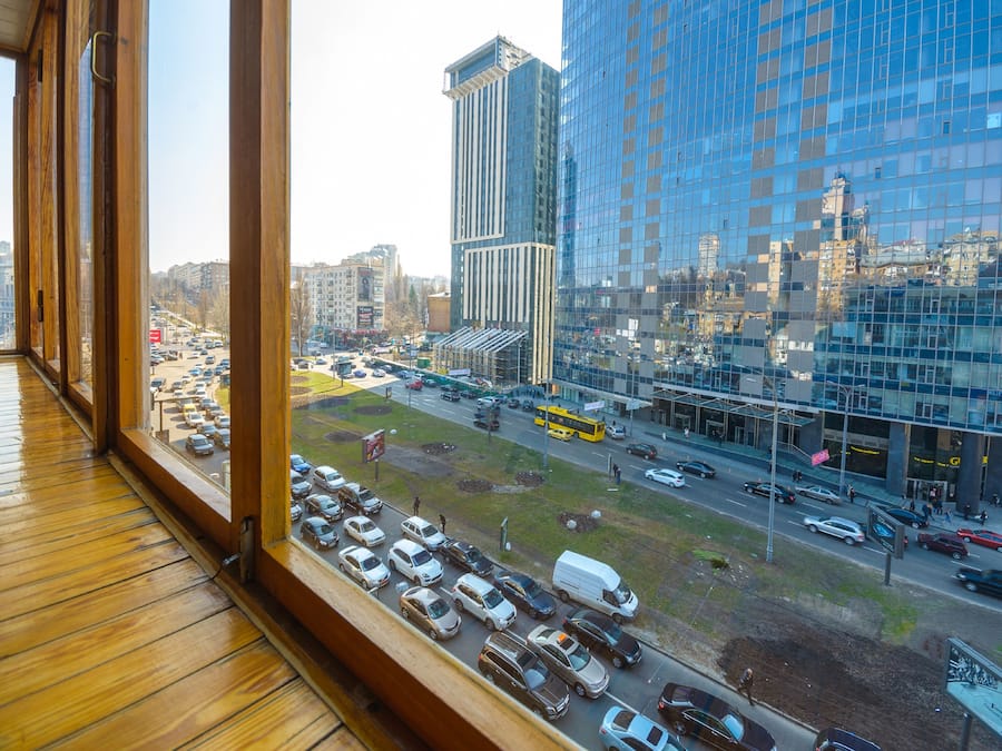 A view from a window overlooking a busy urban street with tall buildings, parked cars, and a yellow bus.
