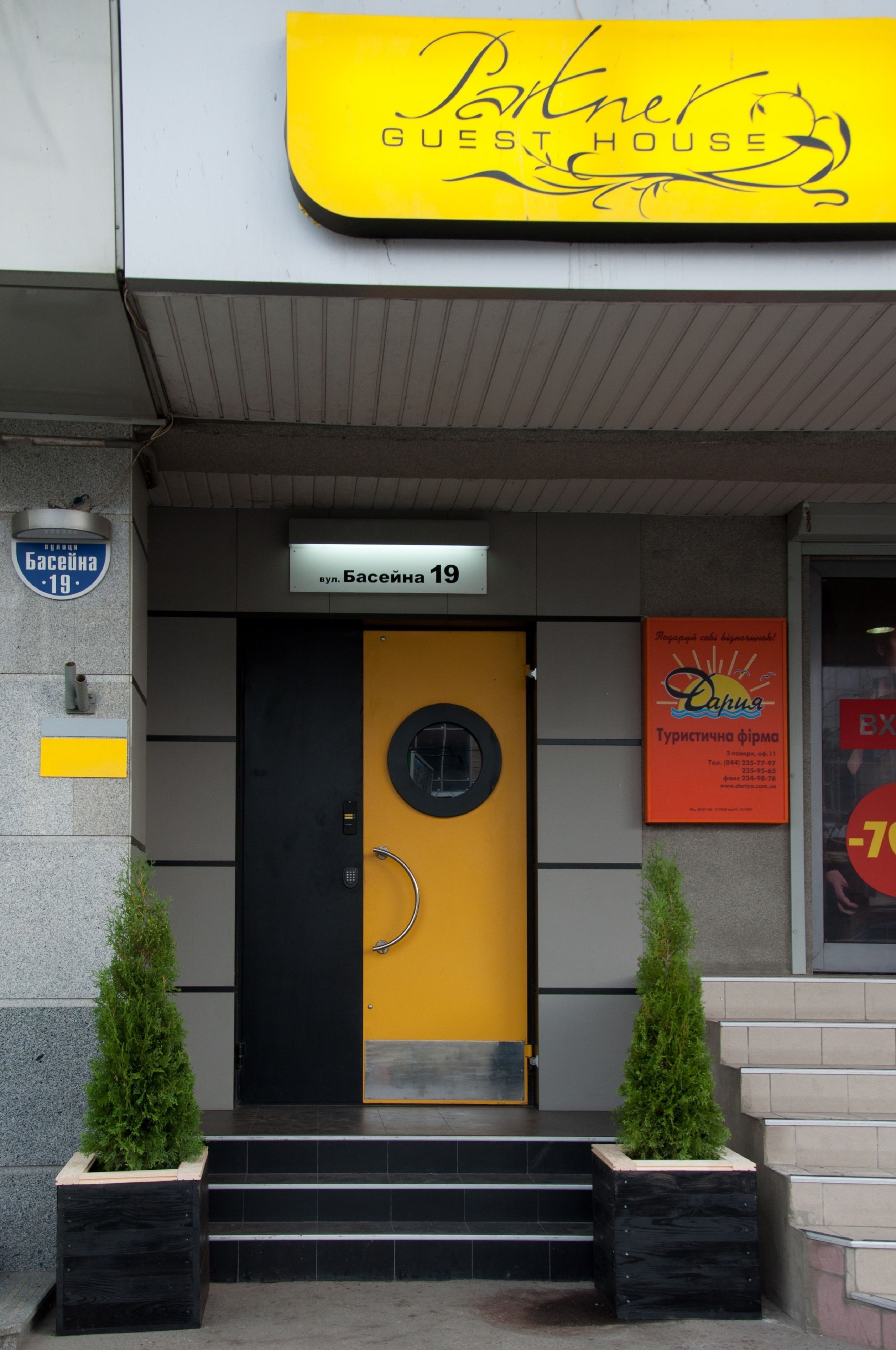 L’entrée d’un hôtel, avec une porte jaune, une enseigne indiquant « Basieina 19 » et, au-dessus, une autre enseigne indiquant « Maison d’hôtes partenaire ».