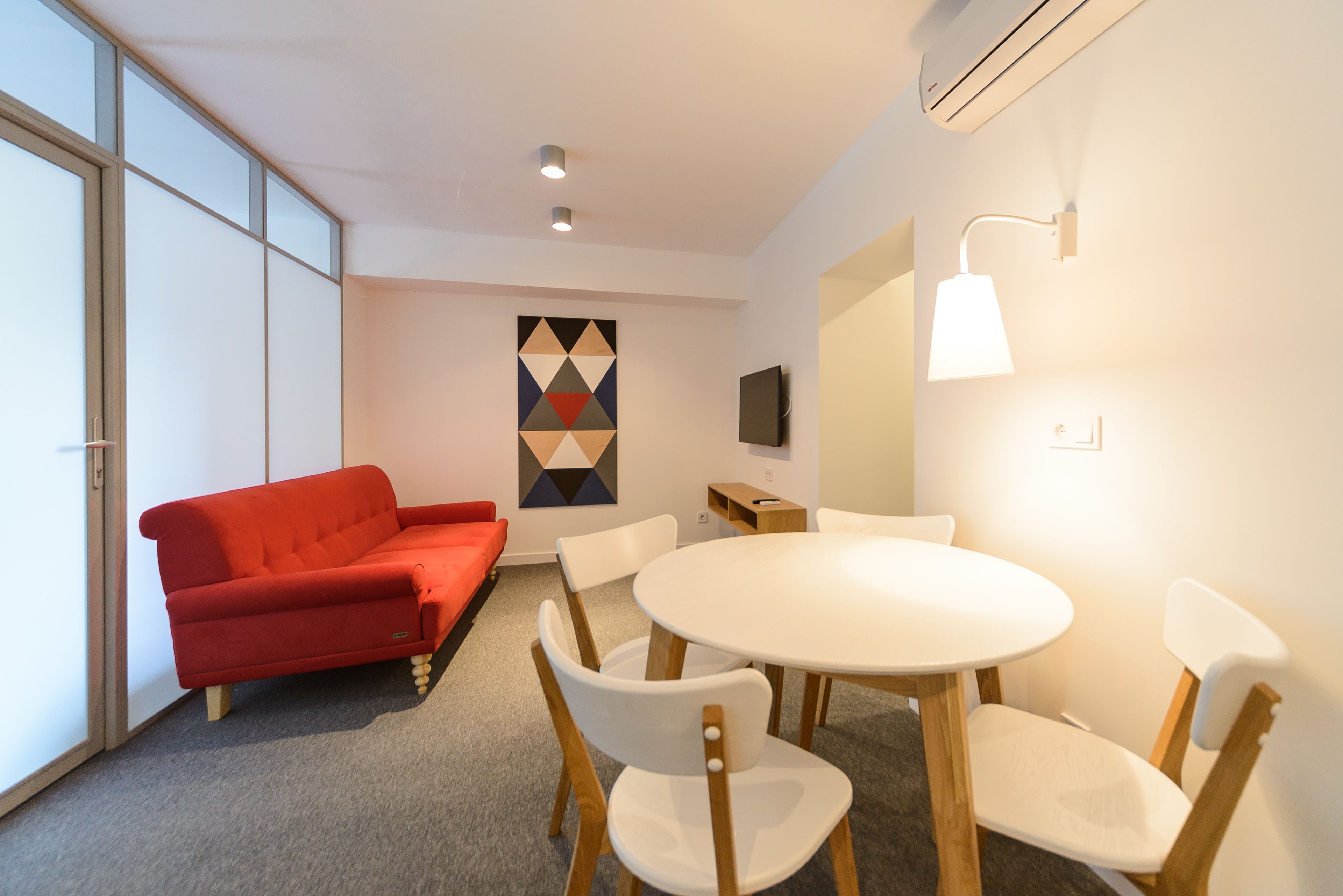 A modern room with a red sofa, a round table with chairs, a geometric wall art piece, and a mounted TV.