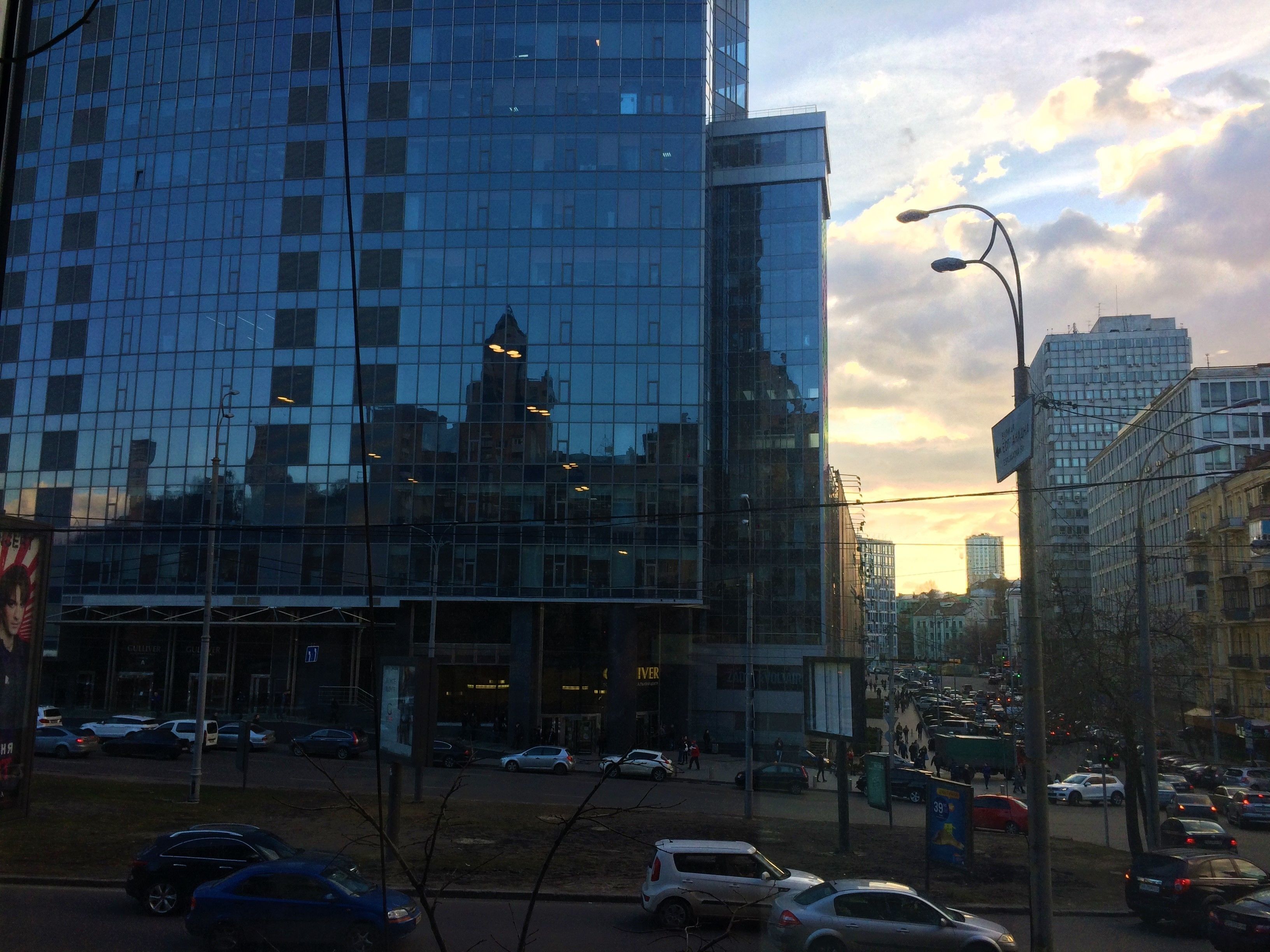 Um edifício moderno de vidro refletindo edifícios circundantes e uma rua movimentada com carros e pedestres.