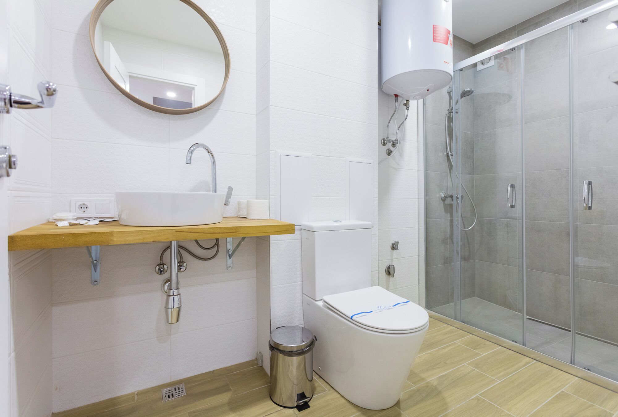 A modern bathroom with a wooden vanity, a white sink, a toilet, a trash can, a shower enclosure, and a round mirror.