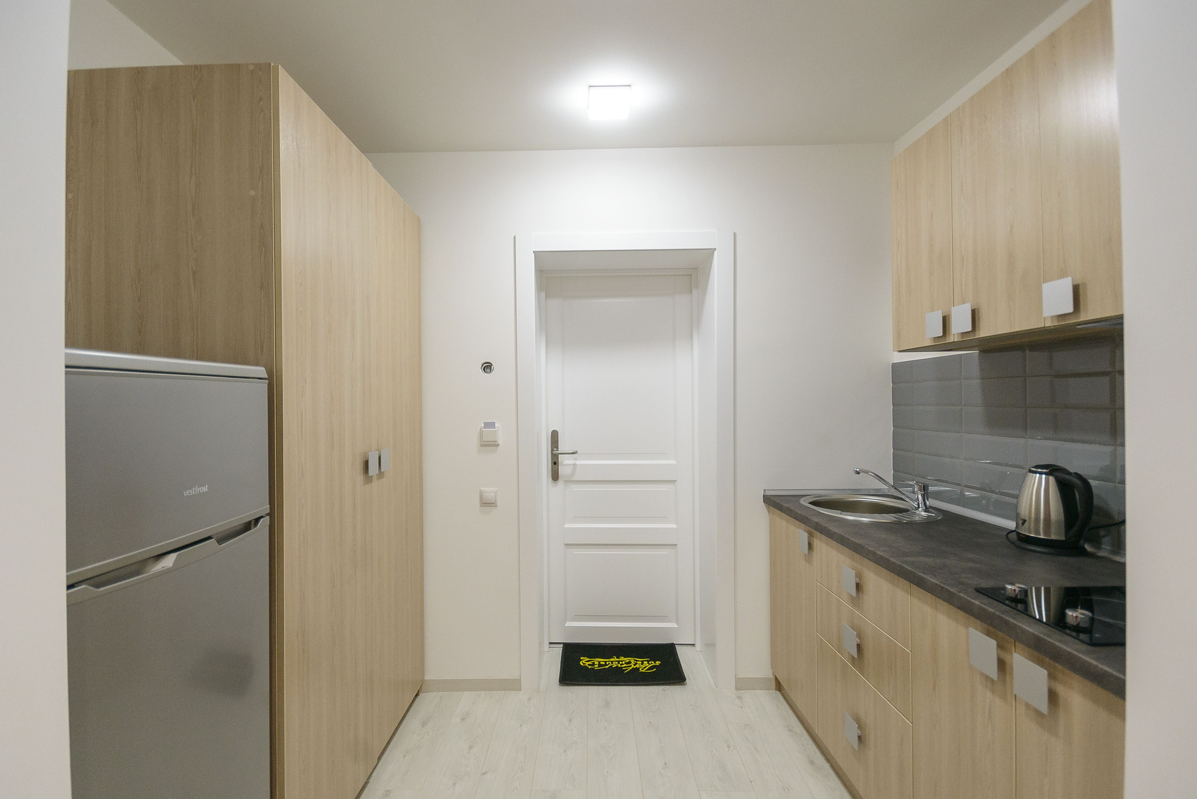 A compact kitchen with wooden cabinets, a stainless steel refrigerator, a sink, and a kettle.