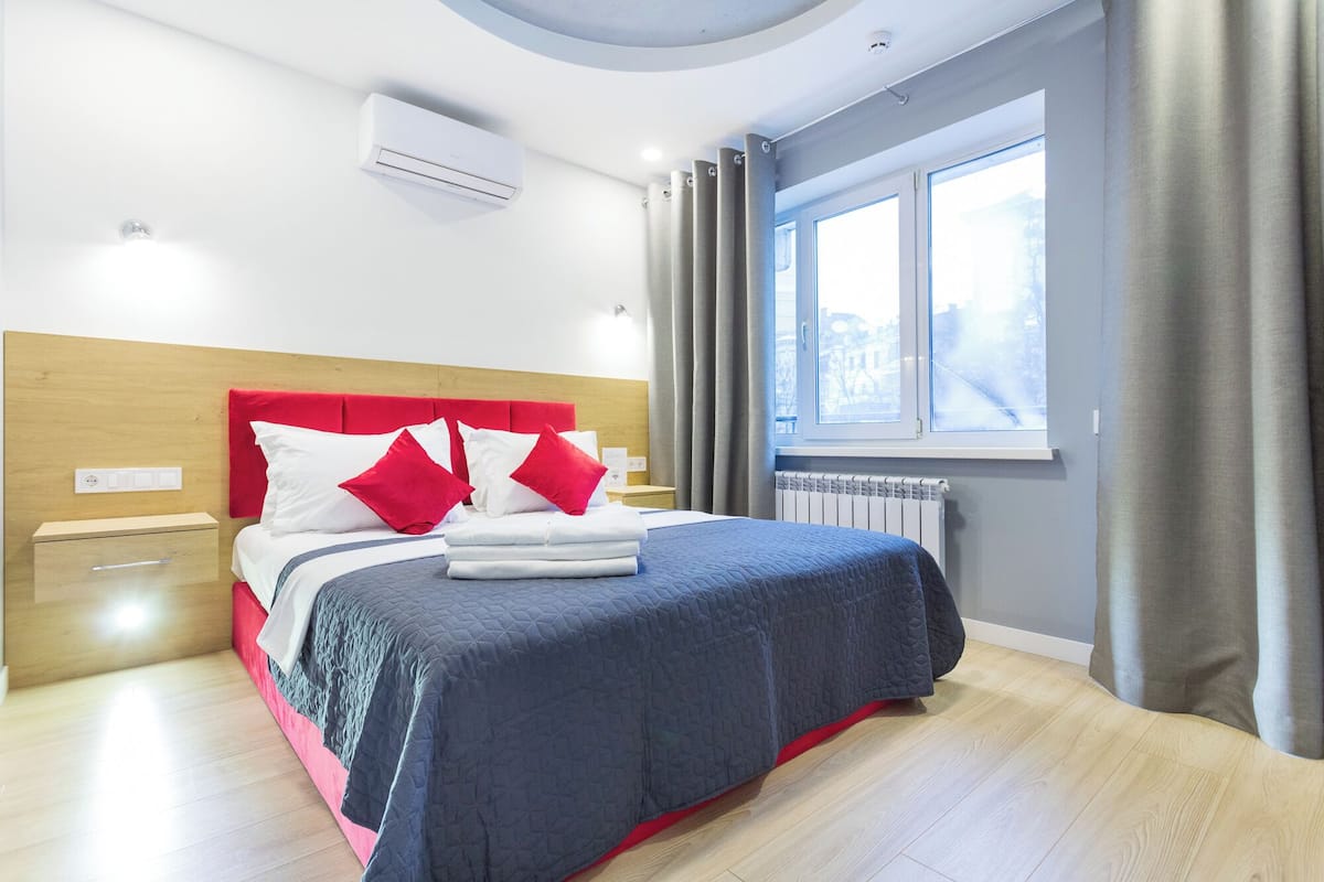 A modern hotel room with a bed, a bedside table, a nightstand, a window with curtains, and an air conditioning unit.