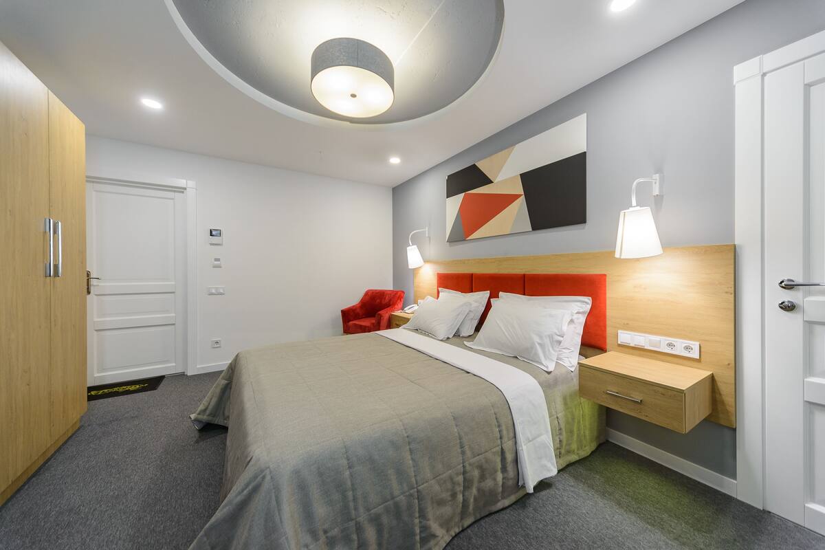 A modern hotel room with a bed, a nightstand, a red chair, and a wall-mounted lamp.