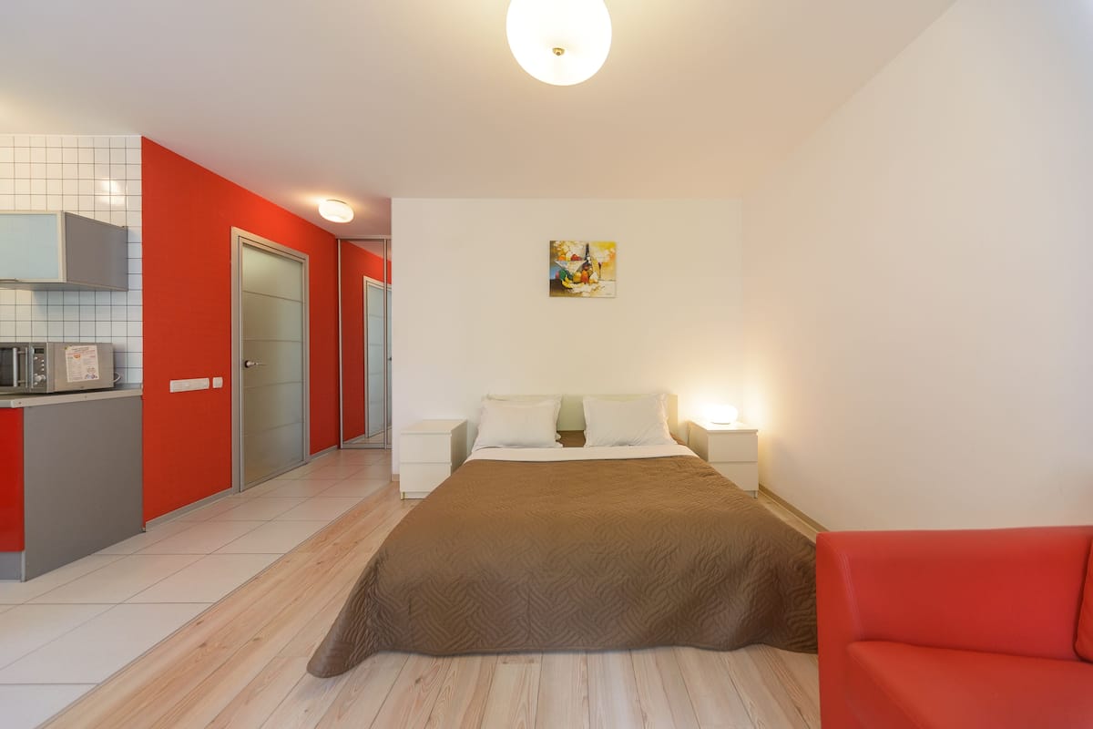 A modern hotel room with a kitchen area, a bed with a brown blanket, and a red sofa.
