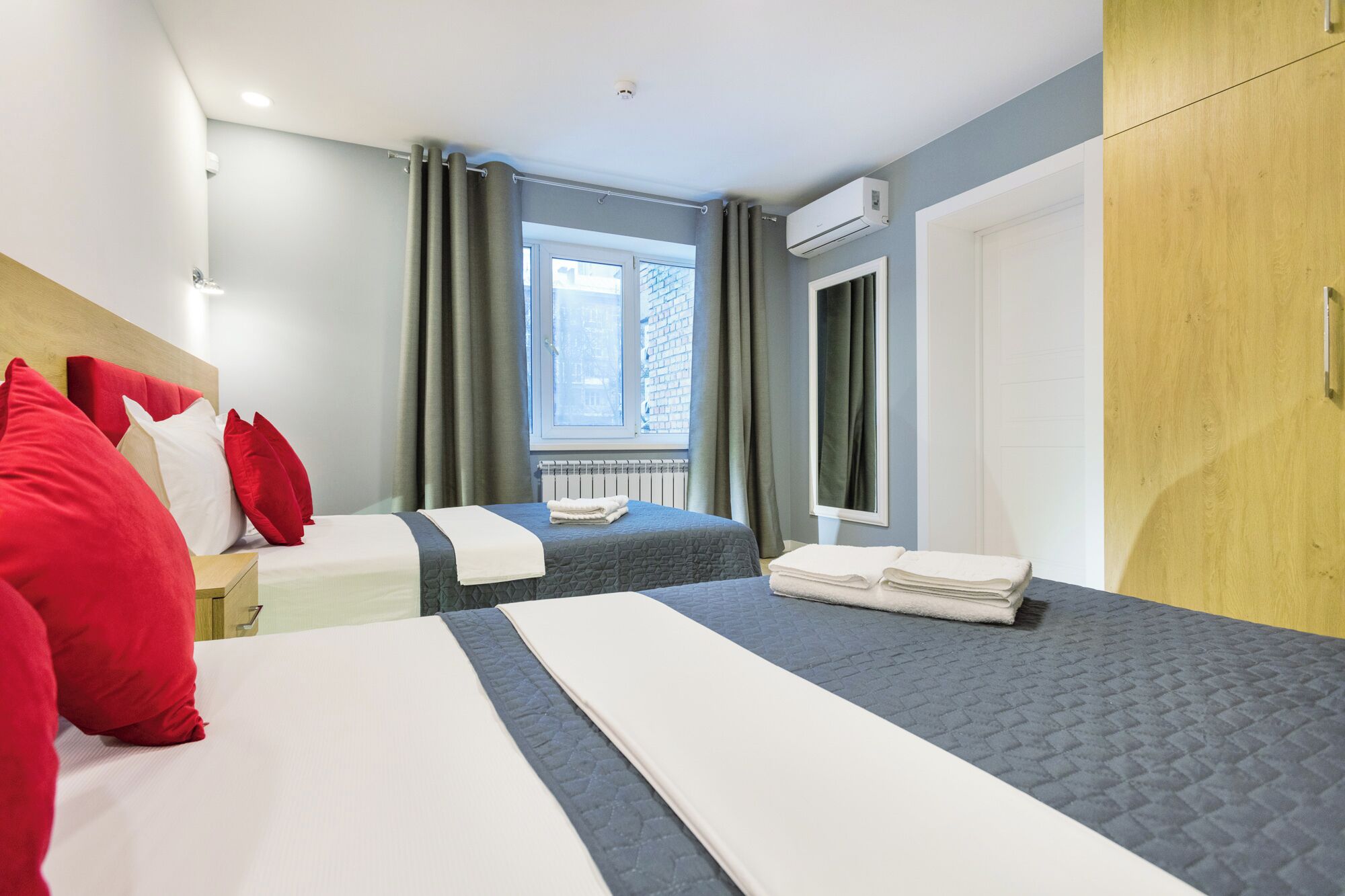 Une chambre d’hôtel avec deux lits, une literie grise et blanche, des oreillers rouges, une fenêtre donnant sur un bâtiment en briques, un appareil de climatisation et une armoire en bois.