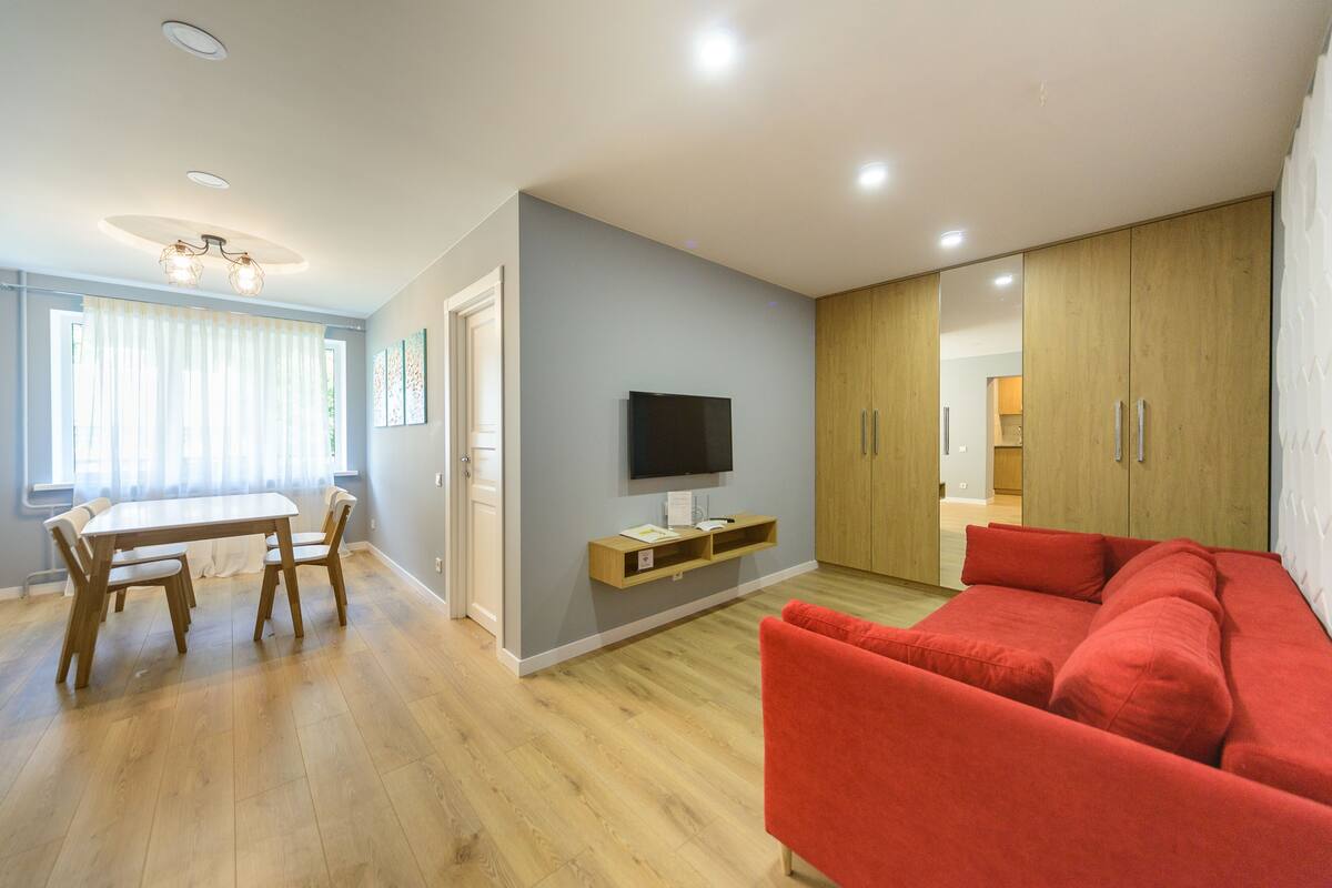 A modern living room with a red sofa, wooden dining table, and a TV mounted on the wall.