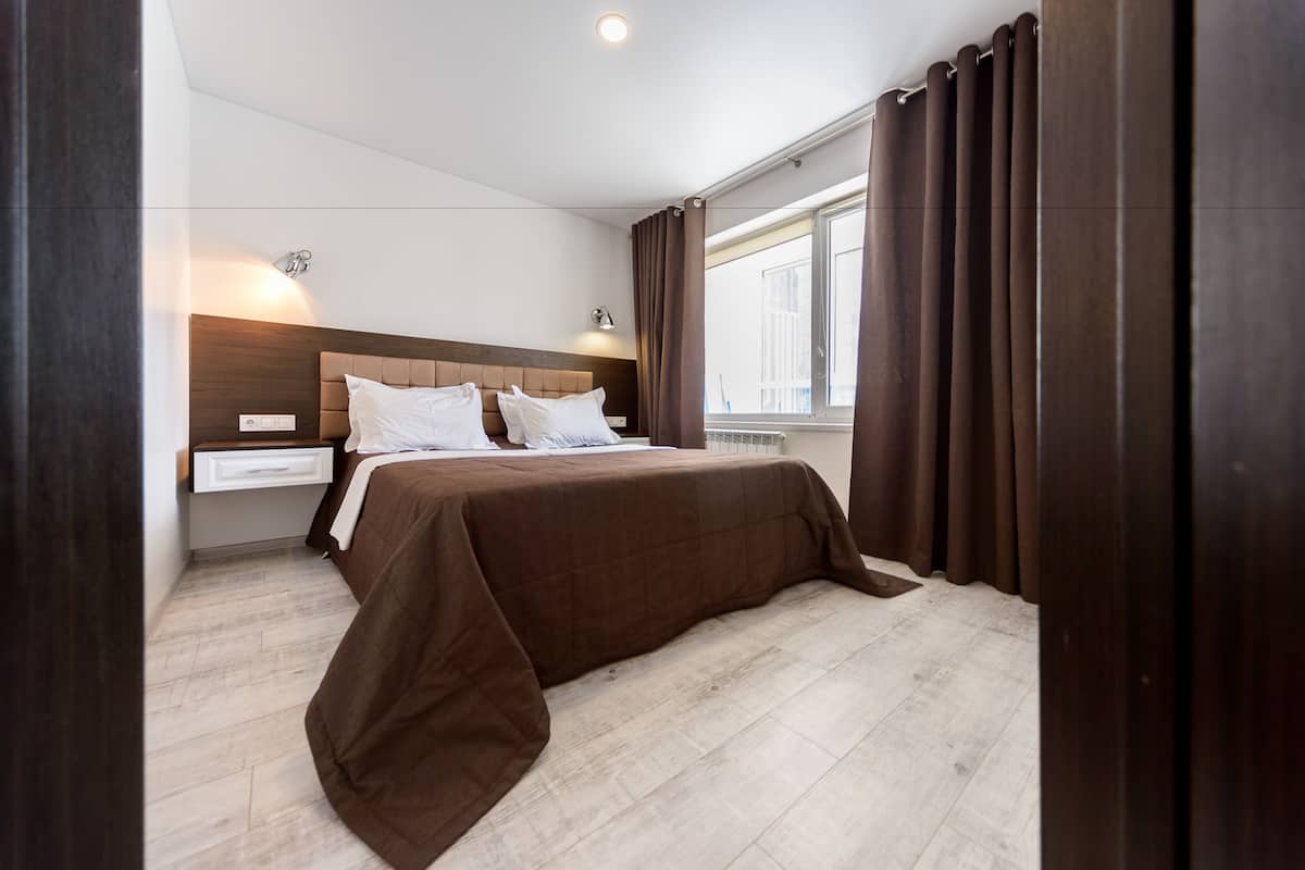 A modern bedroom with a large bed, a nightstand, a wall-mounted light, and a window with curtains.