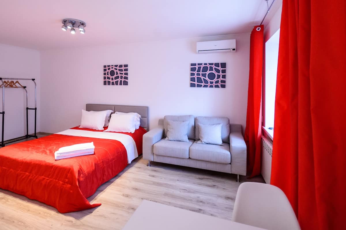 A hotel room with a bed, a sofa, a table, and a window with red curtains.