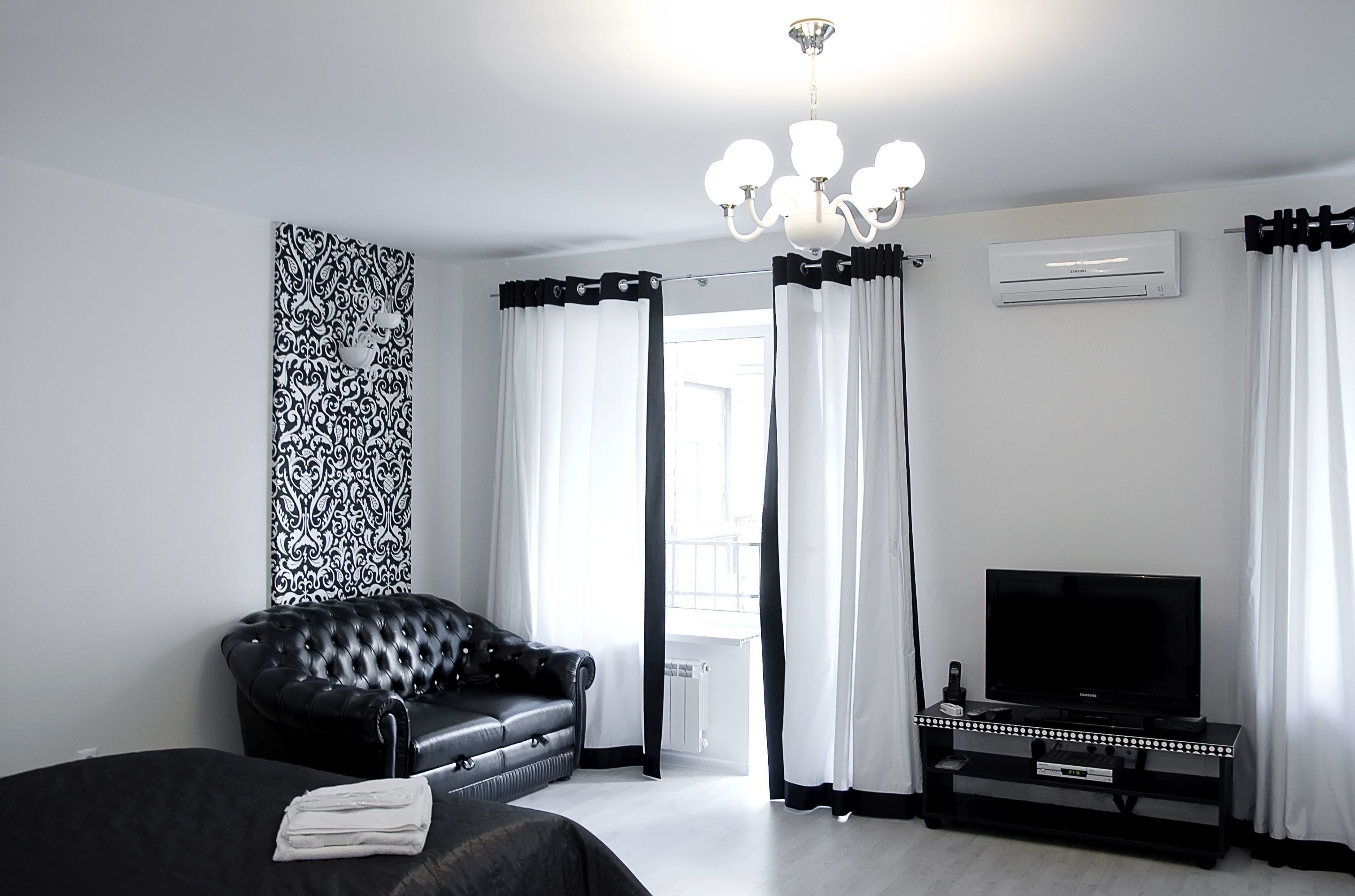 Une chambre d’hôtel moderne avec un canapé en cuir noir, un téléviseur à écran plat posé sur un meuble noir, un lustre et une grande fenêtre ornée de rideaux blancs.