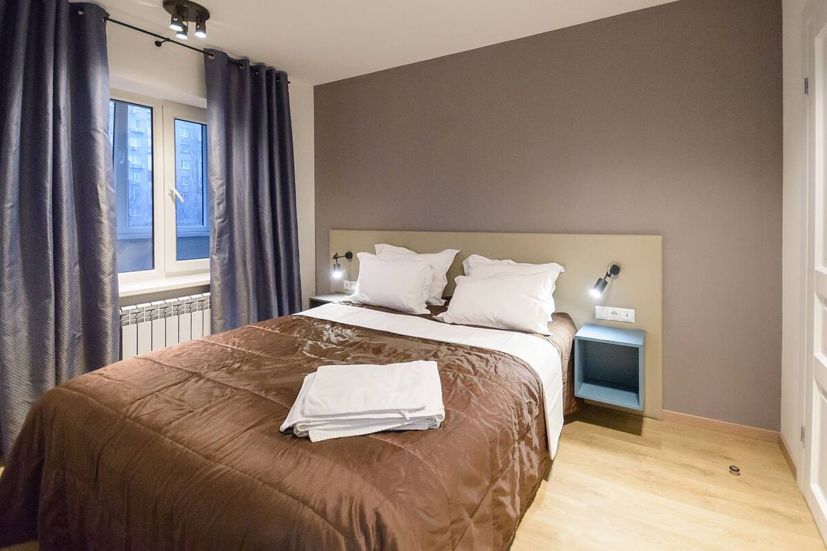A bedroom with a large bed, a nightstand, a window with curtains, and a view of the outside.