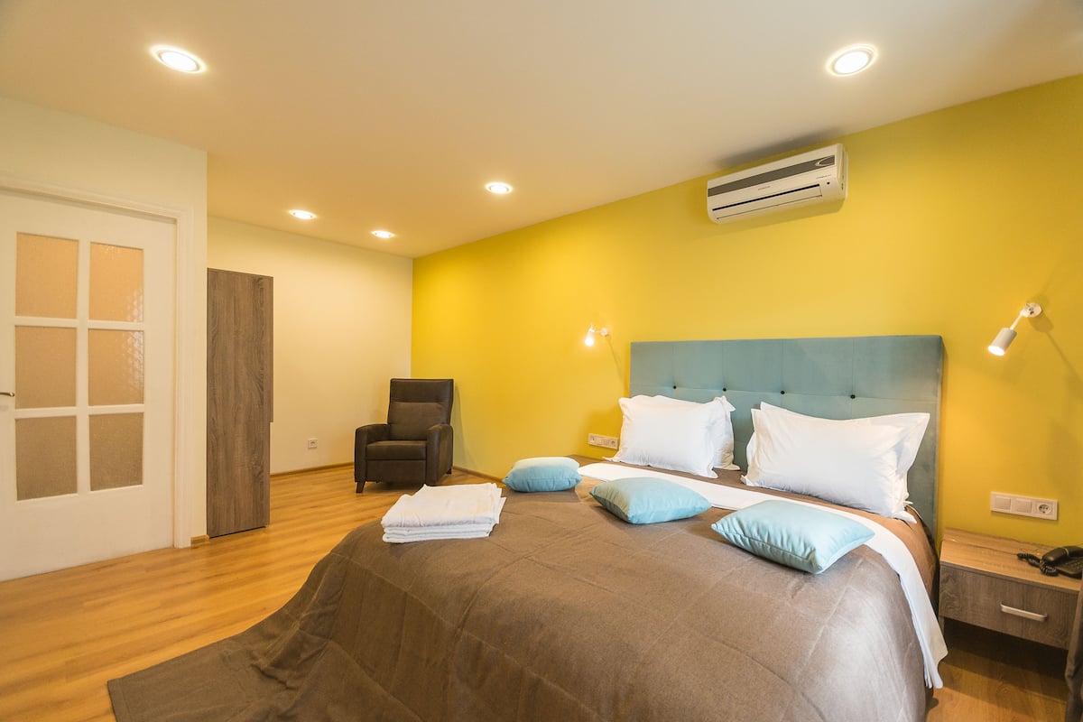 A bedroom with a bed, a chair, a bedside table, an air conditioner, and a door.