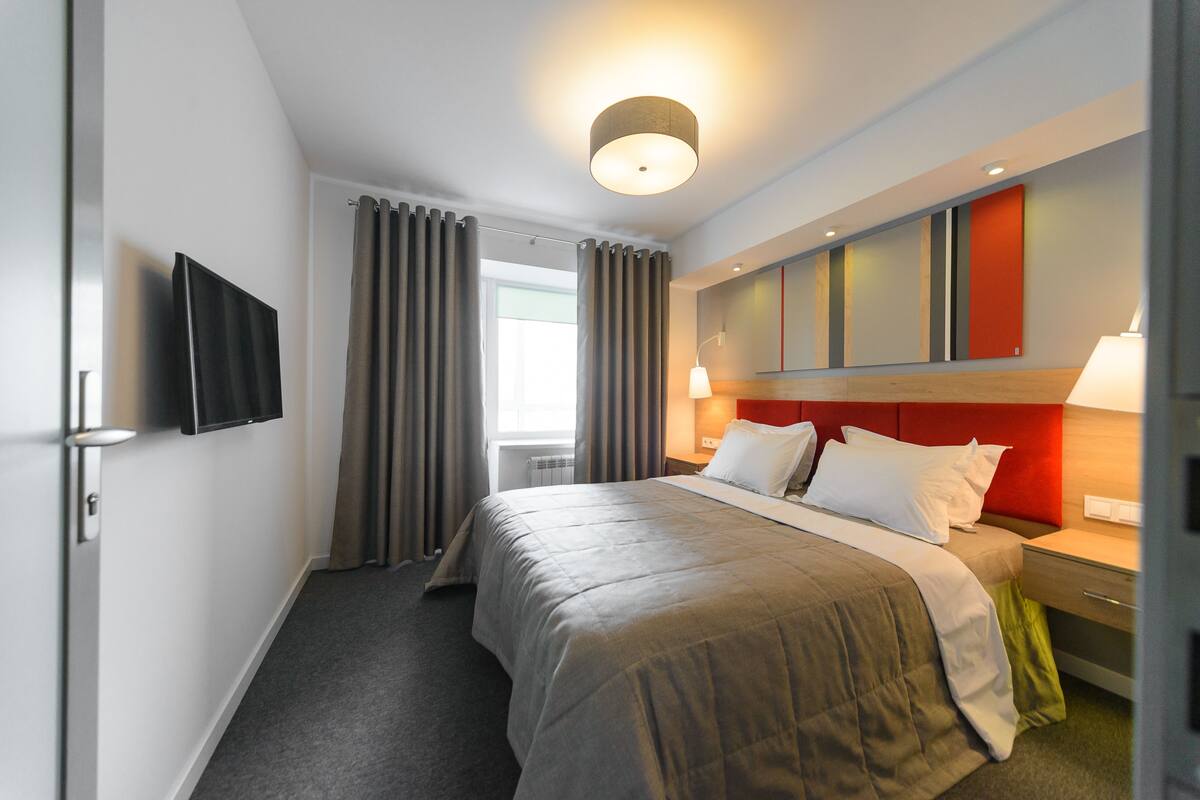 A modern hotel room with a large bed, a flat-screen TV mounted on the wall, a bedside table with a lamp, and a window with curtains.