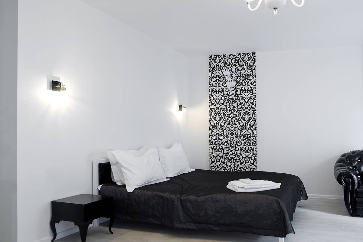 A modern bedroom with a black bed, white pillows, a black and white patterned wall, a small bedside table, and a black leather chair.