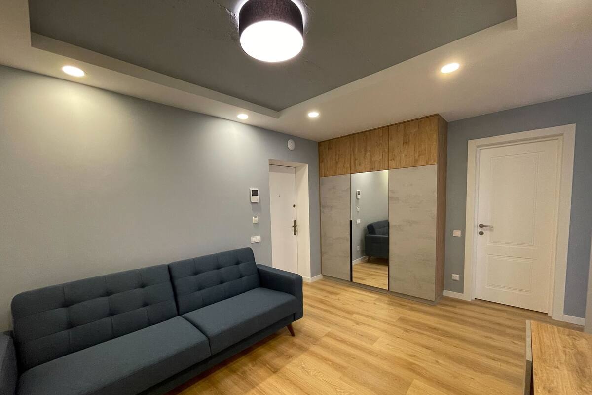 A modern living room with a grey sofa, wooden flooring, and recessed ceiling lights.